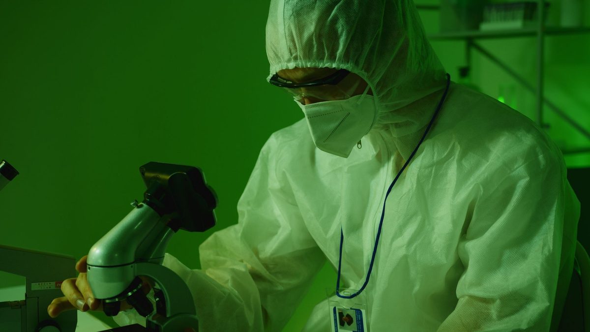 Profesional de laboratorio con traje de protección observando por el microscopio bajo luz verde