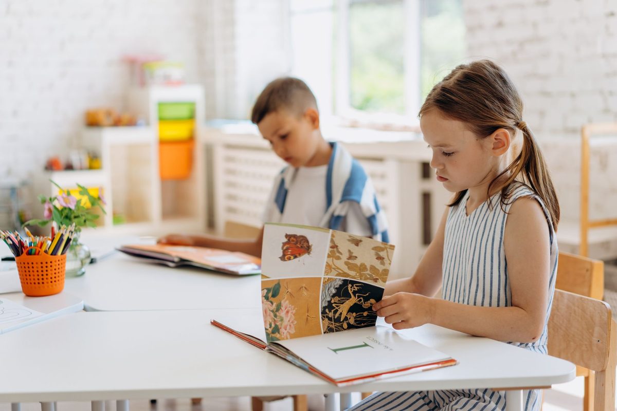 Dos niños leyendo libros ilustrados en una mesa junto a la ventana